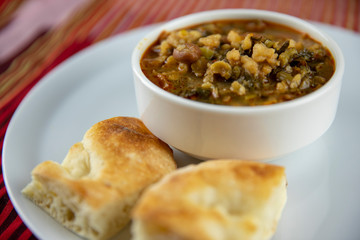 A bowl of hearty soup with chunks of vegetables and grains, served with pieces of crusty bread on a white plate, accompanied by a wooden utensil, set against a vibrant red and gold striped background.