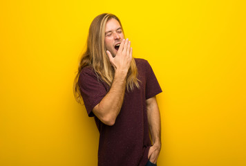 Blond man with long hair over yellow wall yawning and covering wide open mouth with hand