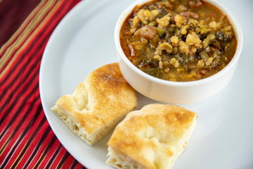 A bowl of hearty soup with chunks of vegetables and grains, served with pieces of crusty bread on a white plate, accompanied by a wooden utensil, set against a vibrant red and gold striped background.