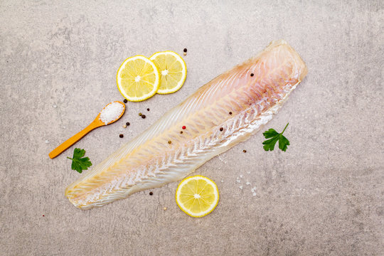 Fresh raw hake fillet on a stone background. With lemon, parsley, salt in wooden spoon and peppers mix. Top view