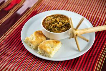 A bowl of hearty soup with chunks of vegetables and grains, served with pieces of crusty bread on a white plate, accompanied by a wooden utensil, set against a vibrant red and gold striped background.