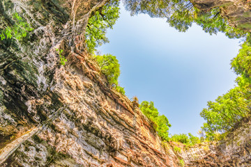 Fototapeta premium Bottom view of rocks and trees in the Melissani caves with a blue sky background. Kefalonia, Greece.