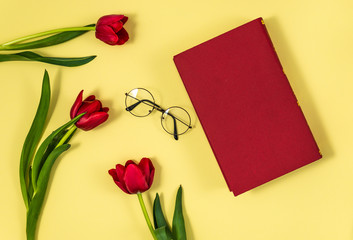Red tulips with red book and glasses on yellow background , flat lay top view