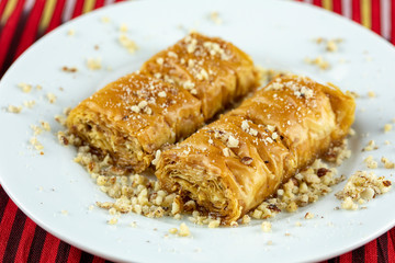 Two pieces of golden, flaky baklava on a white plate, garnished with crushed nuts, set against a vibrant red and gold striped background.