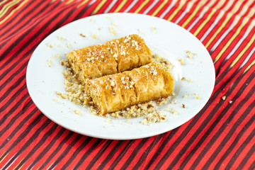 Two pieces of golden, flaky baklava on a white plate, garnished with crushed nuts, set against a vibrant red and gold striped background.