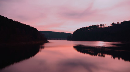 A pink pastel-colored sky and the silhouette of the forest reflect in a calm lake. Oker Dam, Harz mountains, Lower Saxony, Germany.