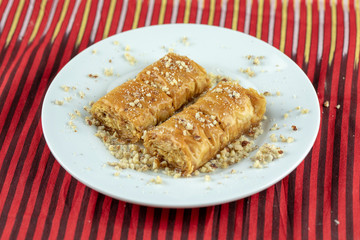 Two pieces of golden, flaky baklava on a white plate, garnished with crushed nuts, set against a vibrant red and gold striped background.