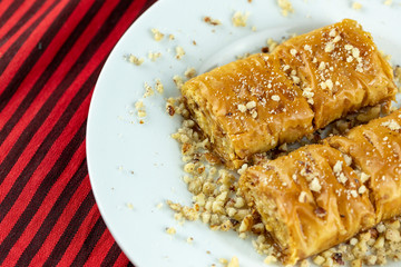 Two pieces of golden, flaky baklava on a white plate, garnished with crushed nuts, set against a vibrant red and gold striped background.