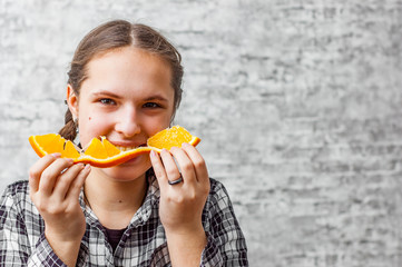portrait of young teenager brunette girl with long hair eat orange fruit on gray wall background...