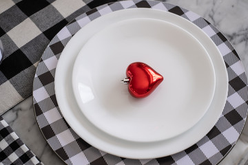 A red shiny valentnes heart sits on a plate on a dining room table with checkerboard table cloths and napkins