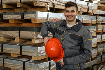 Happy male worker wearing orange helmet and protective suit holding his hardhat on metal stock....