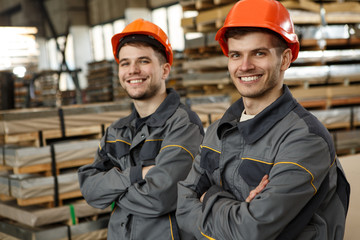 Two young colleagues in gray uniforms and orange helmets standing together with folded arms, smiling, looking at camera and posing on metal stock. Teamwork and collaboration concept.