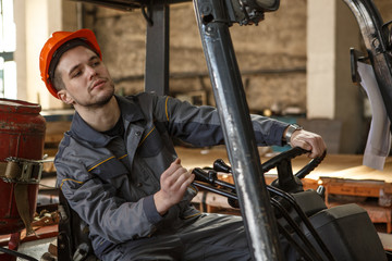 Thorougful worker of metal stock in orange hardhat and protective suit in process of driving forklift. Concentrated toung man looking carefully while transferring material.