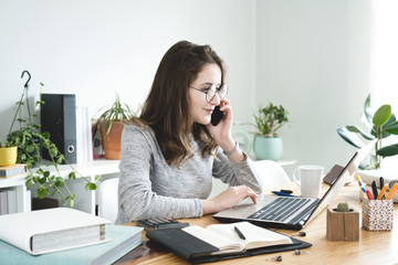 Obraz premium Young business woman working on laptop and talking mobile phone in office.