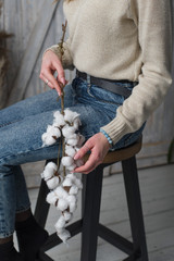 a girl in a sweater with an aquamarine bracelet holds a branch of cotton on her arm, the girl sits on a chair (vertically)