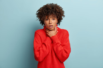 Dissatisfied black woman with curly hair, keeps hands on thore throat, suffers of suffocation, cant breath well, has stressful look, wears oversized red sweater, isolated over blue background.