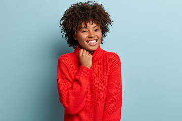 Close up shot of gentle woman with satisfied expression, appealing appearance, keeps hand on collar of red sweater, smiles broadly, has white perfect teeth, isolated over blue background. Emotions
