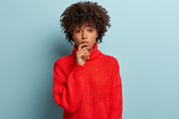 Shot of attractive sad woman has upset frustrated look, touches lips with finger, has thoughtful expression, dark skin, wears red jumper, isolated over blue background. Ethnicity and feminity concept