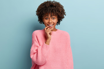 Joyous attractive woman with satisfied expression, keeps fore finger on lips, smiles broadly, wears...