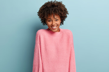 Happy joyful woman with pleased expression, expresses good emotions, wears pink sweater, isolated over blue background. Optimistic lady being in high spirit, satisfied to recieve compliment.