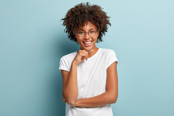 Portrait of cheerful black woman with Afro hairstyle, smiles gently, imagines something pleasant, wears round glasses and t shirt models against blue background. Emotions and human expressions concept
