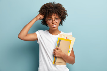 Puzzled female student scratches head, tries to learn material, has crisp hair, carries spiral notebook and papers, forgets answer on question, isolated over blue background. Studying concept