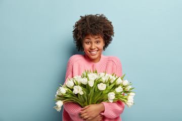 Optimisitc young lady with satisfied expression carries beautiful white flowers closely to herself, dressed in oversized pink sweater, celebrates holiday during spring, models over blue studio wall