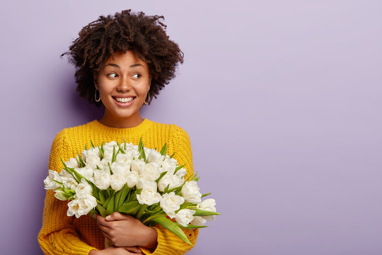 Studio shot of cheerful woman with curly hairstyle, gazes aside, has white teeth, holds bouquet of tulips, models against purple background, likes spring time. Mothers Day and holidays concept