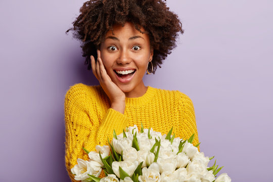 Cropped image of overjoyed woman with curly hair, touches cheek, wears yellow knitted jumper, enjoys spring time, surprised to recieve white tulips, isolated over purple wall. Bunch of flowers for you