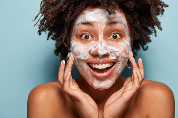 Happy lady wets face with foam, stands in studio with bare shoulders, touches cheeks, has Afro...