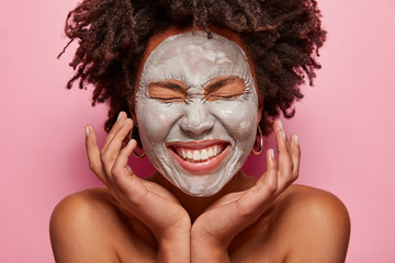 Face care procedure concept. Gentle beautiful woman touches face, smiles broadly, shows white teeth, has clay mask on face, poses half nude, isolated over pink background. Anti aging concept