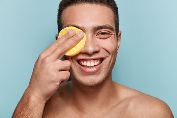 Pleased young guy holds yellow pad on eye, smiles gladfully, removes cosmetic cream, wants to have perfect smooth healthy skin, clean body shows white even teeth, poses over blue background.