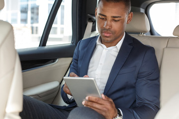 Young businessman sitting on back seat in car browsing digital tablet concentrated