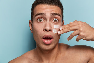 Headshot of stupefied man applies moisturizer cream, has bugged eyes, short hairstyle, uses anti aging lotion, has bare shoulders, isolated over blue background. Skin care and masculinity concept - obrazy, fototapety, plakaty