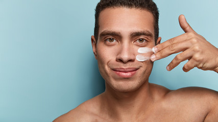 Close up shot of attractive man with healthy skin, applies cream for anti wrinkle or anti aging,...