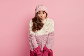 Amazed beautiful European woman with astonished facial expression, wonders on shocking revelation, has wavy hair, wears headgear and loose warm sweater, poses in studio against pink background