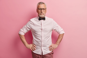 Self confident wrinkled man keeps hands on waist, wears shirt with black bowtie, looks directly at camera, isolated over pink background, has serious expression. People, age and pension concept