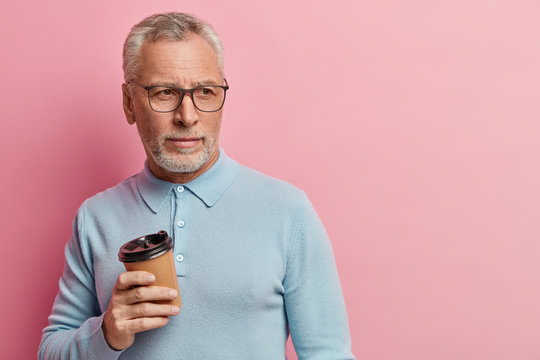 Photo of pensive bearded man dressed in casual blue jumper, holds takeaway coffee, looks aside, has rest, thinks about future plans, isolated over rosy studio wall with empty space for your slogan