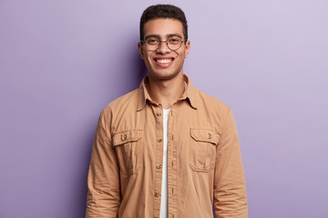Handsome smiling guy wears spectacles and beige shirt, looks at camera with satisfaction, enjoys getting new job position, isolated over purple background. Youth and positive emotions concept