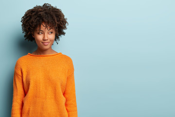 Image of mixed race woman with cunning sly expression, looks curliously aside, has intention to implement plan in life, has Afro hairstyle, wears orange sweater, models over blue background.