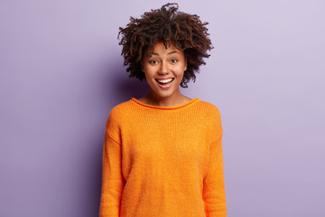 Indoor shot of attractive woman with cheerful expression, has toothy smile, dressed in orange jumper, expresses joy, isolated over purple background. Human facial expressions and emotions concept