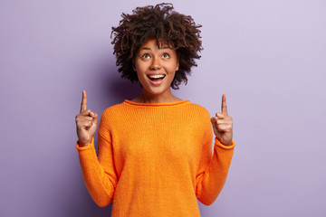 Positive young good looking woman with Afro hairstyle, indicates with both index fingers, wears...
