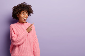 Positive emotional African American woman with curly hairstyle, wears pink sweater, indicates with...