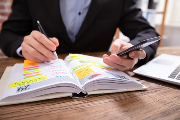 Businessman Holding Cellphone Writing Schedule In Diary With Pen