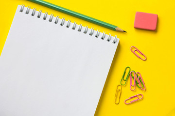 Notepad on yellow background, green pencil, paper clips and eraser top view
