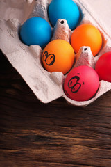 Colorful Easter eggs with painted face in egg carton box on wooden background. Space for text