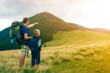Fototapeta na wymiar Father and son with backpacks hiking together in summer mountains. Back view of dad and child holding hands on landscape mountain view. Active lifestyle, family relations, weekend activity concept