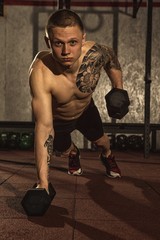 Fototapeta na wymiar Vertical portrait of a young handsome shirtless muscular man doing pushups and lifting dumbbells, while working out at crossfit gym studio. Muscles, energy, achievement, active lifestyle concept