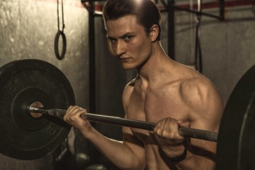 Obraz na płótnie Canvas Shot of a young attractive man exercising shirtless at the gym, looking to the camera fiercely. Handsome athletic man lifting barbell. Workout, agility, strengthening concept