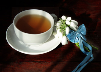 A white cup with green tea on a saucer and a small bouquet of spring white snowdrops, tied with a ribbon. Rustic still life